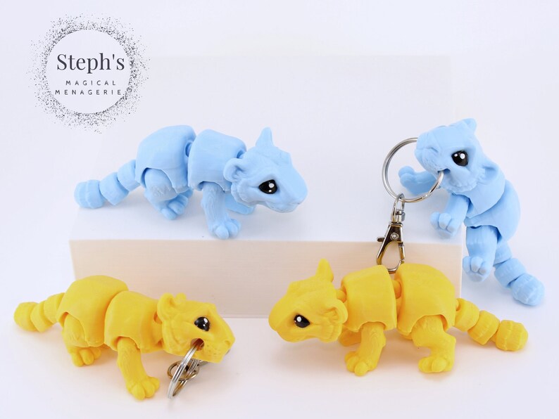 May include: Four articulated animal keychains, two are yellow and two are light blue. The keychains are posed in a playful manner. Steph's Magical Menagerie is printed on the background.