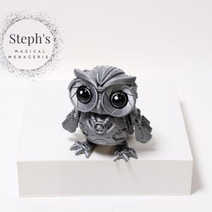 May include: A gray, stylized owl figurine with large, expressive eyes and a mechanical aesthetic. The owl is perched on a white block, with the text "Steph's MAGICAL MENAGERIE" in a circular design above it.