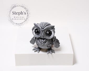 Può includere: Una statuetta di gufo grigio stilizzata con grandi occhi espressivi e un'estetica meccanica. Il gufo è posizionato su un blocco bianco, con il testo "Steph's MAGICAL MENAGERIE" in un design circolare sopra.