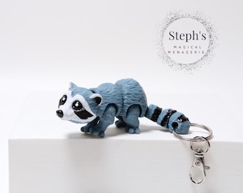 Puede incluir: Un llavero de mapache azul y negro con un cierre giratorio plateado. El mapache está en posición de caminar con la cola enroscada. El llavero está etiquetado como "Steph's Magical Menagerie".