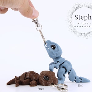 Puede incluir: Dos figuras de dinosaurio articuladas para llavero, una marrón y otra azul acero. El dinosaurio azul está unido a un llavero plateado y está de pie. El dinosaurio marrón está tumbado. El texto "Steph's Magical Menagerie" es visible en el fondo.