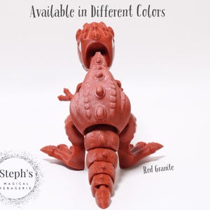 May include: Red granite dinosaur toy with articulated limbs. The dinosaur is sitting on a white surface. Available in different colours.