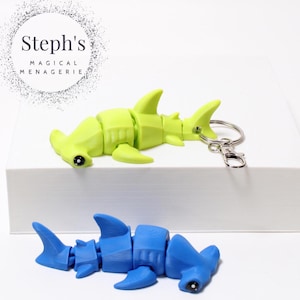 May include: Two articulated hammerhead shark keychains. One is lime green, the other is blue. Both have black eyes and are on a white surface. The green shark has a silver keychain attached. The text "Steph's MAGICAL MENAGERIE" is in a circle in the background.
