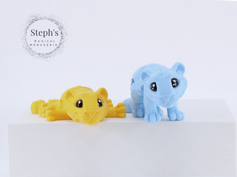 May include: Two small toy tigers, one yellow and one light blue, with black eyes. The tigers are posed in a playful manner.