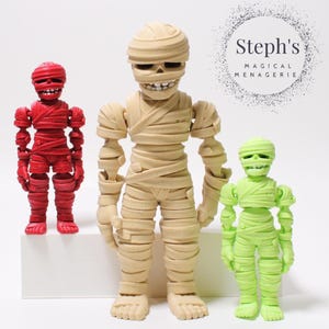 May include: Three articulated mummy figures in red, tan, and green. The mummies have detailed wrappings and skeletal faces. The figures are standing on a white surface. The text "Steph's MAGICAL MENAGERIE" is in a circle in the background.