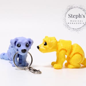 May include: Two dog-shaped keyrings. One is light purple and holds a silver keyring in its mouth. The other is yellow. Both have large black eyes and are posed on a white surface. The text "Steph's MAGICAL MENAGERIE" is in a circle in the background.