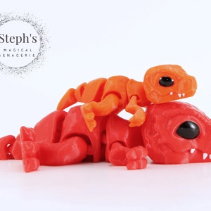 May include: A red and orange articulated dinosaur toy with black eyes and white teeth. A smaller orange dinosaur is perched on top of the larger one. The image includes the text "Steph's Magical Menagerie".