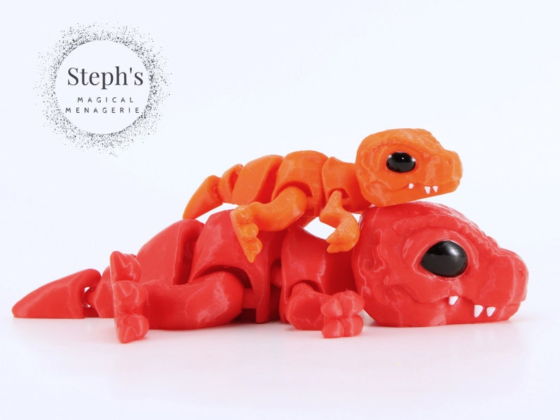 May include: A red and orange articulated dinosaur toy with black eyes and white teeth. A smaller orange dinosaur is perched on top of the larger one. The image includes the text "Steph's Magical Menagerie".