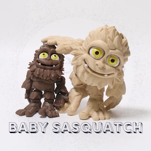 Puede incluir: Dos figuras articuladas de Sasquatch con grandes ojos amarillos y pelaje texturizado. Una figura es de color marrón oscuro y la otra es de color beige claro. El texto BABY SASQUATCH está impreso en letras blancas y negritas abajo.