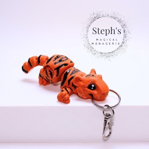 Pode incluir: Chaveiro de tigre laranja e preto com um anel e um fecho prateados. O tigre é feito de argila polimérica e tem um estilo de desenho animado. O chaveiro é etiquetado como "Steph's Magical Menagerie".
