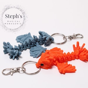 Może przedstawiać: Dwa kolorowe breloczki z smokiem, wydrukowane w 3D. Jeden jest niebieski, a drugi pomarańczowy. Oba mają srebrny kółko do kluczy.