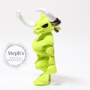 May include: A lime green figurine of a bull-like creature with white horns and black eyes. The figure has a muscular build and is posed in a standing position. The figure has a gold-colored ring on its ear. The text "Steph's MAGICAL MENAGERIE" is in a circle on the left.