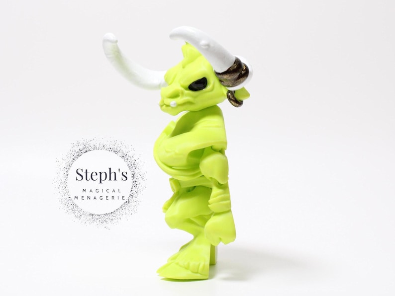 May include: A lime green figurine of a bull-like creature with white horns and black eyes. The figure has a muscular build and is posed in a standing position. The figure has a gold-colored ring on its ear. The text "Steph's MAGICAL MENAGERIE" is in a circle on the left.
