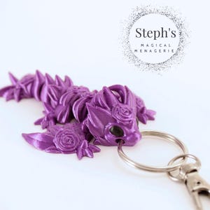 May include: Purple dragon keychain with a rose design. The dragon is curled up with its wings spread out. The keychain is attached to a silver key ring with a silver clasp.