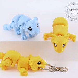 Könnte beinhalten: Drei bewegliche Tiger-Figuren, eine blaue, zwei gelbe, wobei ein gelber Tiger an einem silbernen Schlüsselanhänger befestigt ist. Die Tiger haben schwarze Augen und sind in einer verspielten, hockenden Position dargestellt. Der Text "Steph's Magical Menagerie" ist im Hintergrund sichtbar.