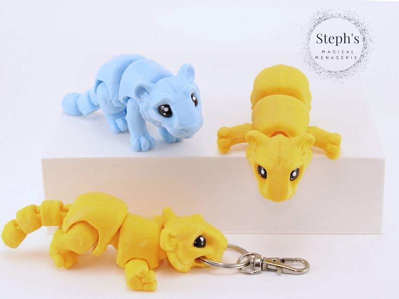 May include: Three articulated tiger figurines, one blue, two yellow, with one yellow tiger attached to a silver key chain. The tigers have black eyes and are posed in a playful, crouching position. The text "Steph's Magical Menagerie" is visible in the background.