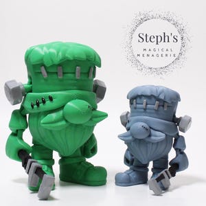 Puede incluir: Dos figuras caprichosas con temática de Frankenstein. Una es verde, la otra azul grisáceo. Ambas tienen detalles de pernos, rasgos parecidos a barbas y sostienen una herramienta en forma de martillo. El texto "Steph's MAGICAL MENAGERIE" está en un círculo.