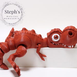 May include: A red, 3D printed dinosaur toy with a large, white eye and a toothy grin. The dinosaur is lying on its side on a white surface.