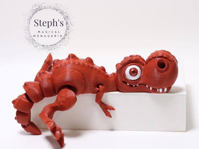 May include: A red, 3D printed dinosaur toy with a large, white eye and a toothy grin. The dinosaur is lying on its side on a white surface.