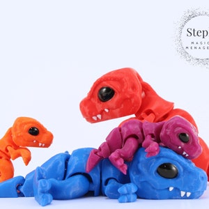 Op de afbeelding: Een verzameling beweegbare drakenfiguren in verschillende kleuren: oranje, rood, blauw en paars. De draken hebben grote zwarte ogen en zijn speels gerangschikt. De afbeelding bevat de tekst "Steph's MAGICAL MENAGERIE".