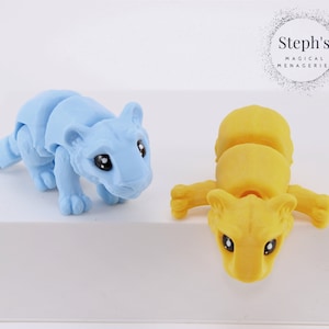 May include: Two articulated toy tigers, one light blue and one yellow, posed on a white surface. Steph's Magical Menagerie is printed on a black circle in the background.