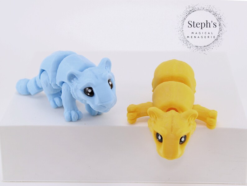 May include: Two articulated toy tigers, one light blue and one yellow, posed on a white surface. Steph's Magical Menagerie is printed on a black circle in the background.