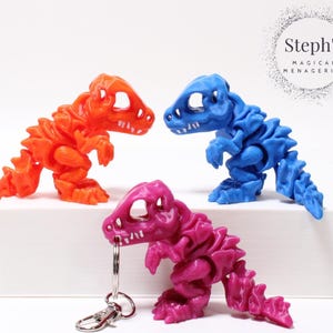 Puede incluir: Tres figuritas de dinosaurios esqueleto, una naranja, una azul y una rosa. El dinosaurio rosa tiene un llavero plateado adjunto.