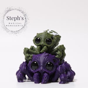 Peut inclure: Figurine fantaisiste représentant une araignée violette avec de grands yeux, portant sur son dos une araignée verte plus petite. L'araignée verte a des marques blanches. Il s'agit probablement d'un objet de collection ou décoratif.
