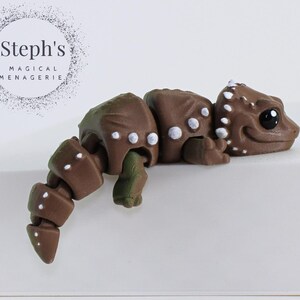 May include: Brown and green articulated toy lizard with white spots and black eyes. The toy is posed in a walking position.