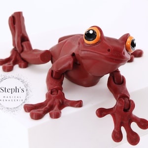 May include: A red articulated frog toy with orange eyes. The frog is posed in a sitting position with its legs outstretched.