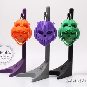 Könnte beinhalten: Drei farbige schädelartige Schlüsselanhänger in Orange, Lila und Grün, jeder mit einem lächelnden Gesicht und einem silbernen Schlüsselring. Sie werden auf Ständern in passenden Farben ausgestellt. Der Text "Steph's Magical Menagerie" befindet sich im Hintergrund.