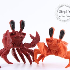 Puede incluir: Dos pequeños cangrejos impresos en 3D, uno rojo y otro naranja, con ojos negros. Los cangrejos están posados como si estuvieran caminando.