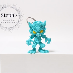 Puede incluir: Una figura de monstruo turquesa con ojos amarillos y un llavero plateado. La figura tiene un diseño detallado y texturizado y se encuentra sobre una superficie blanca. El texto "Steph's MAGICAL MENAGERIE" está en el fondo.