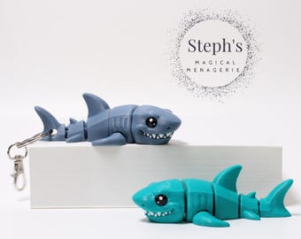 Könnte beinhalten: Zwei bewegliche Hai-Spielzeuge. Eines ist grau und an einem Schlüsselanhänger befestigt, das andere ist türkis. Beide haben schwarze Augen und weiße Zähne. Der Text "Steph's MAGICAL MENAGERIE" befindet sich in einem Kreis im Hintergrund.