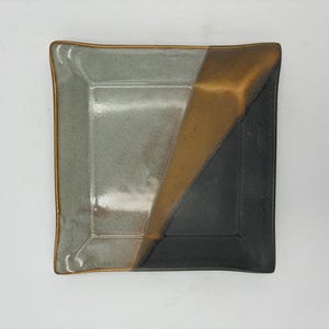 May include: A square ceramic dish featuring a glossy finish with a color-blocked design. The surface is divided into three distinct sections: a light gray area, a metallic bronze diagonal stripe, and a matte black corner section with raised edges.