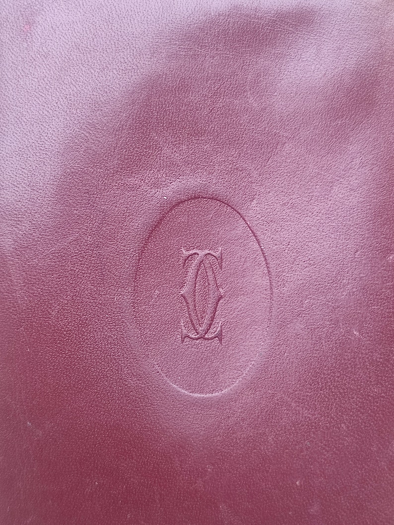 May include: A close-up of a burgundy leather surface with a raised oval-shaped logo. The logo is a stylized letter 'C' with a curved line through the center. The logo is embossed into the leather.