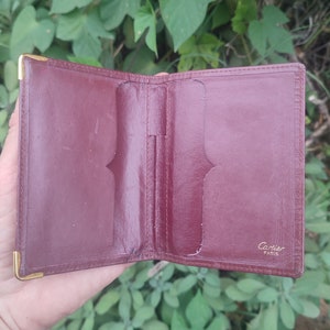 May include: A burgundy leather wallet with gold accents and the words 'Cartier Paris' embossed on the front.