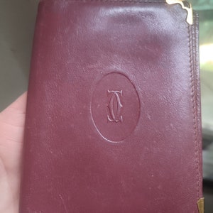 May include: A burgundy leather wallet with gold accents and a circular logo that says 'Cartier'.