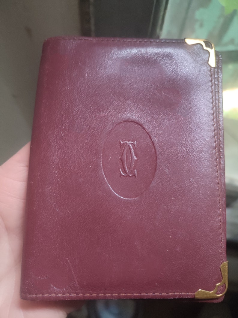 May include: A burgundy leather wallet with gold accents and a circular logo that says 'Cartier'.
