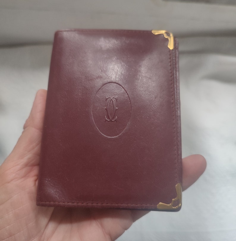 May include: A burgundy leather wallet with gold accents and the Cartier logo embossed on the front.