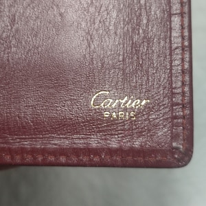 May include: Close-up of a burgundy leather wallet with gold stitching and the words 'Cartier Paris' embossed in gold lettering.