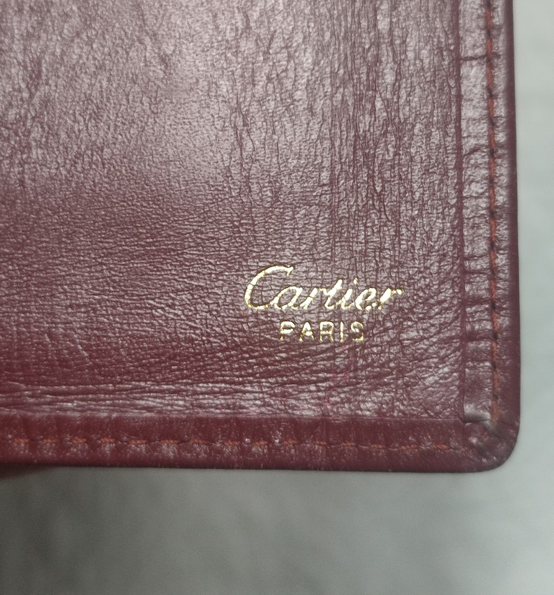 May include: Close-up of a burgundy leather wallet with gold stitching and the words 'Cartier Paris' embossed in gold lettering.