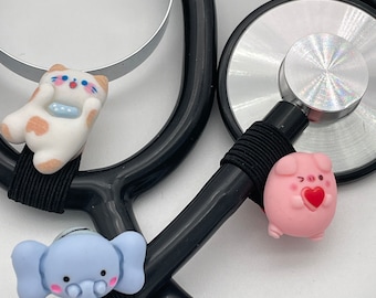 May include: A black stethoscope with three colorful animal charms. The charms are a white cat, a pink pig, and a blue elephant. The charms are attached to the stethoscope with black elastic bands.