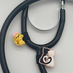 May include: A black stethoscope featuring two decorative charms attached to the tubing. One charm is a yellow dog with a pink bow, and the other charm depicts a black cat and a white cat sitting together with their tails forming a heart shape.