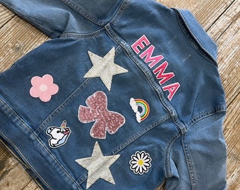 Könnte beinhalten: Eine blaue Jeansjacke mit verschiedenen Patches, darunter eine rosa Blume, ein silberner Stern, eine rosa Schleife, ein Regenbogen, eine weiße Gänseblümchen und ein Einhorn. Die Jacke hat den Namen "EMMA" in rosa auf der Rückseite gestickt.