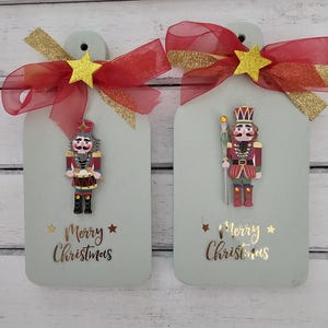May include: Two wooden Christmas decorations featuring a nutcracker design. Each decoration has a red and gold ribbon with a gold star. The decorations display the text "Merry Christmas" in gold lettering.