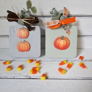 May include: Two wooden signs with a fall theme. Each sign is painted a light blue-green color and features a pumpkin. One sign has two smaller pumpkins and the other has one larger pumpkin. The signs are decorated with brown and orange ribbon and green leaves. Scattered around the signs are orange and white candy corn.