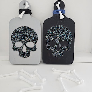 May include: Two wooden cutting boards with glitter skull designs. The boards are painted gray and black. White plastic bones are scattered around the boards.