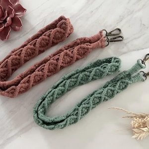 Könnte beinhalten: Zwei Makramee-Taschenriemen in Staubrosa und Salbeigrün. Jeder Riemen hat ein gewebtes Design und Metallverschlussbefestigungen. Die Riemen werden auf einer weißen Marmoroberfläche mit einer Sukkulente und Trockenblumen im Hintergrund präsentiert.