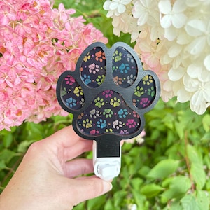 Op de afbeelding: Zwart nachtlampje in de vorm van een pootafdruk met kleurrijke pootafdrukken op een donkere achtergrond. Het nachtlampje heeft een witte stekker. Roze en witte bloemen op de achtergrond.